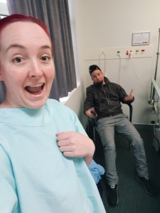 Me taking a giddy selfie in a hospital gown with my boyfriend in the background